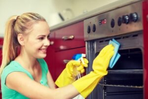 Happy Woman With Bottle Of Spray Cleanser Cleaning Oven At Home Kitchen